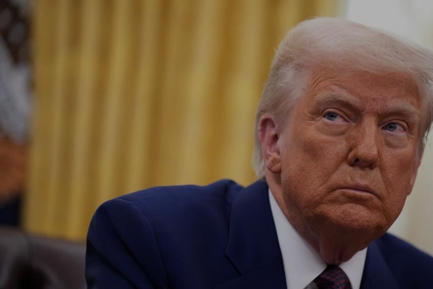 A close-up image of President Trump in the oval office. He has a scowl on his face and is looking to the right. there are yellow curtains in the background.