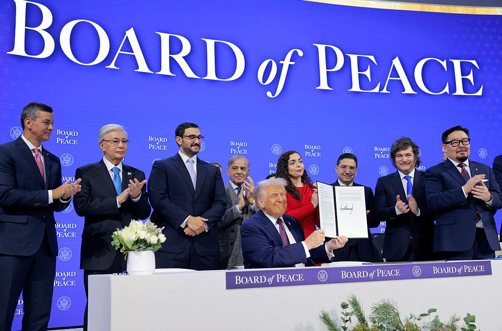 Donald Trump seated at a long table flanked by various officials, with the words "Board of Peace" behind them